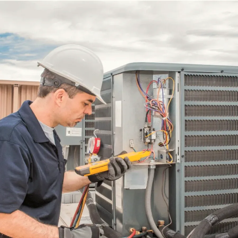 HVAC technician in a hard hat testing an outdoor air conditioner with a multimeter